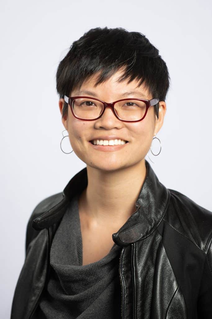 Headshot of an Asian American woman with a pixie cut, wine colored glasses, and silver hoop earrings. She is smiling and wearing a softly draping gray shirt and leather jacket.