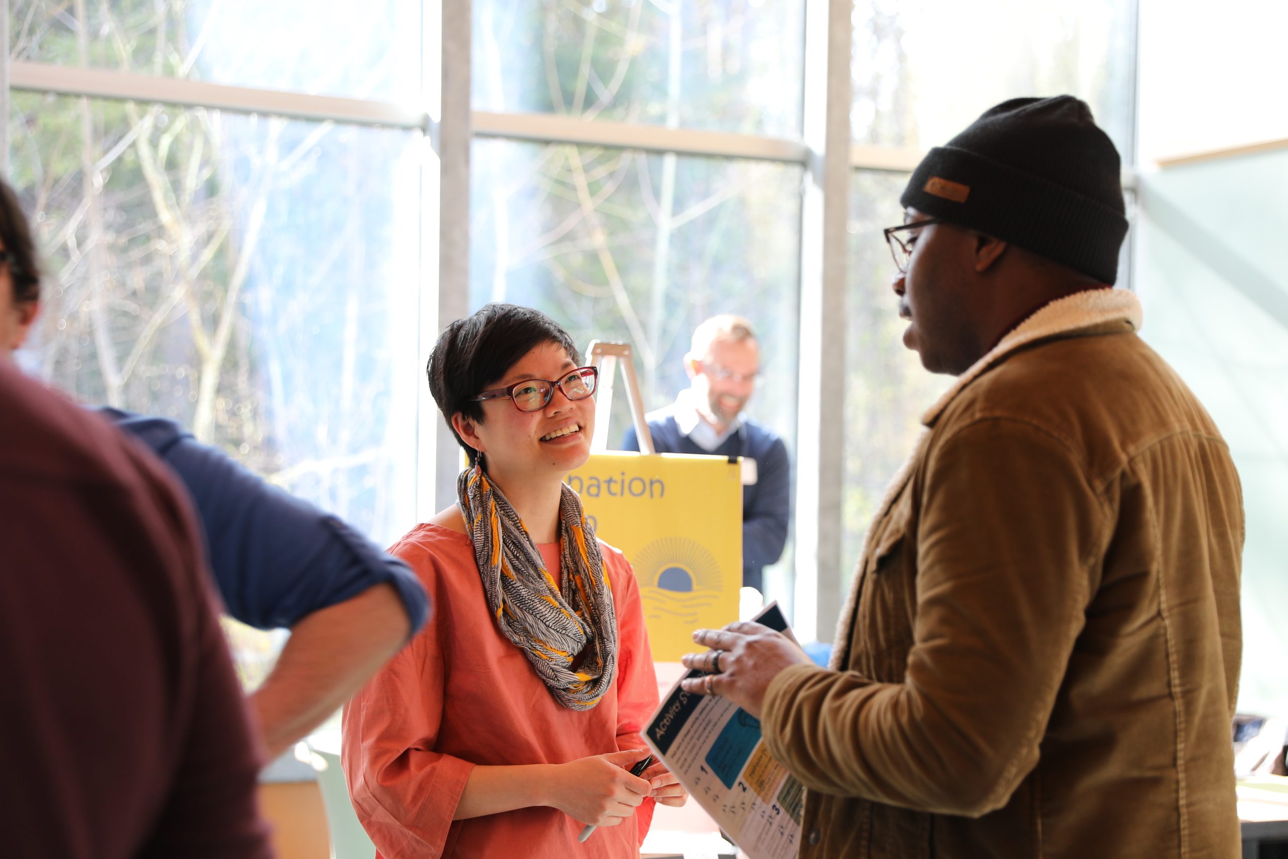 Sophia Xiao-fan Austrins smiles broadly while speaking with a community member at a Nature in Neighborhoods Community Choice Grants workshop. She is wearing a warm salmon colored shirt and patterned scarf and sports a pixie cut and glasses. The community member is a tall Black male wearing a brown jacket, black ski cap, and glasses. He holds colorful handouts. An Imagination Station is visible behind in front of a view out to a forested landscape.